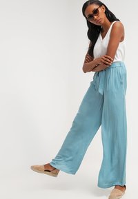 Light blue, wide-leg pants with a tie waistband, pleats, and a soft texture. Paired with a white top and tan slip-on shoes.