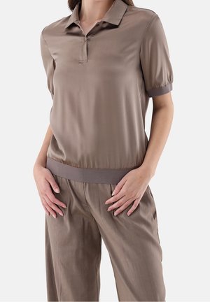 Mujer vistiendo una camisa polo de manga corta color topo con puños y cinturilla acanalados, combinada con pantalones sueltos a juego, de pie con las manos en las caderas.