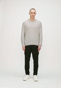 Pulover texturat gri deschis cu guler rotund, purtat cu pantaloni negri și adidași albi, stând pe un fundal alb simplu.