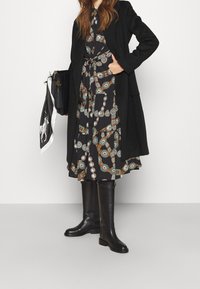 Svart lång kappa över en mönstrad klänning med cirkelmönster i teal och brunt, tillsammans med knähöga svarta stövlar och en handväska.