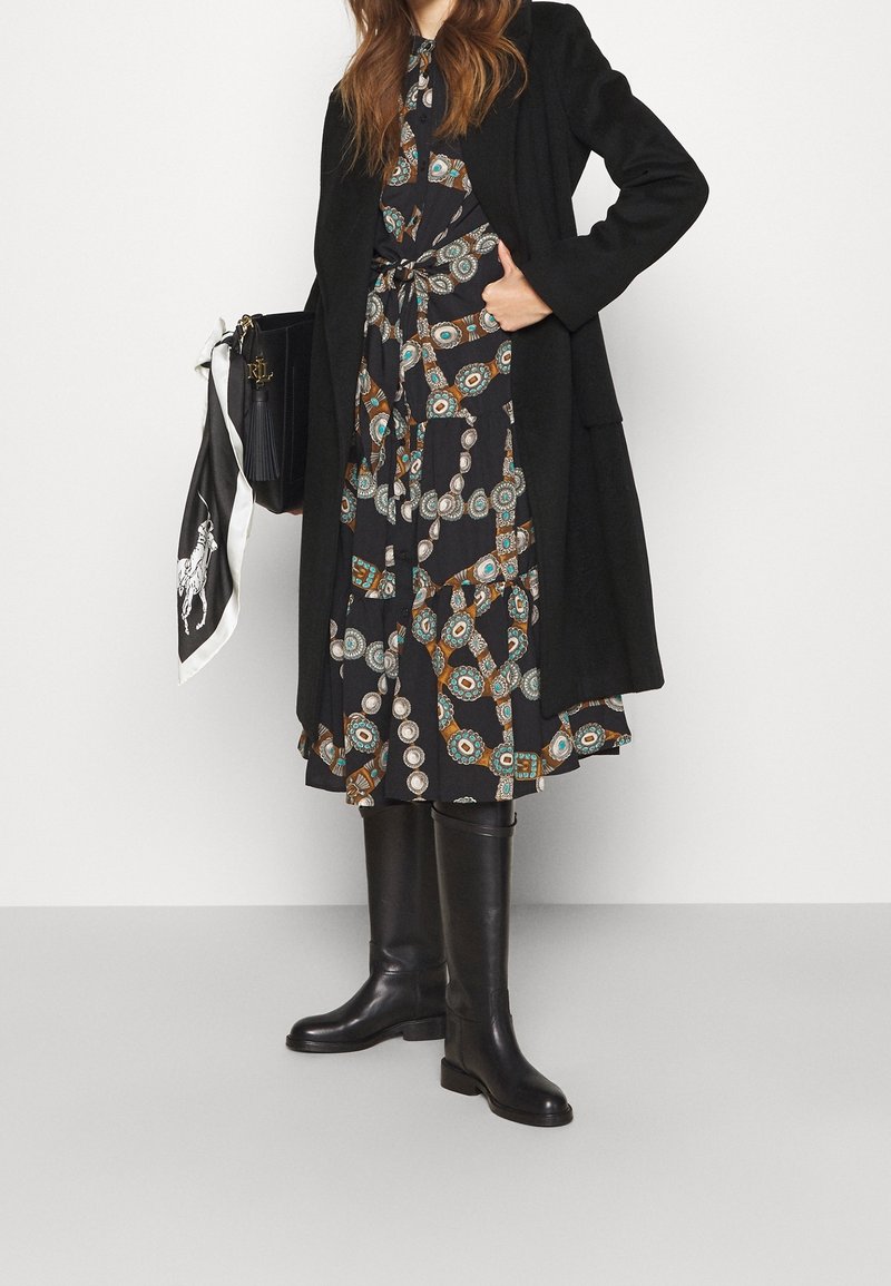 Svart lång kappa över en mönstrad klänning med cirkelmönster i teal och brunt, tillsammans med knähöga svarta stövlar och en handväska.