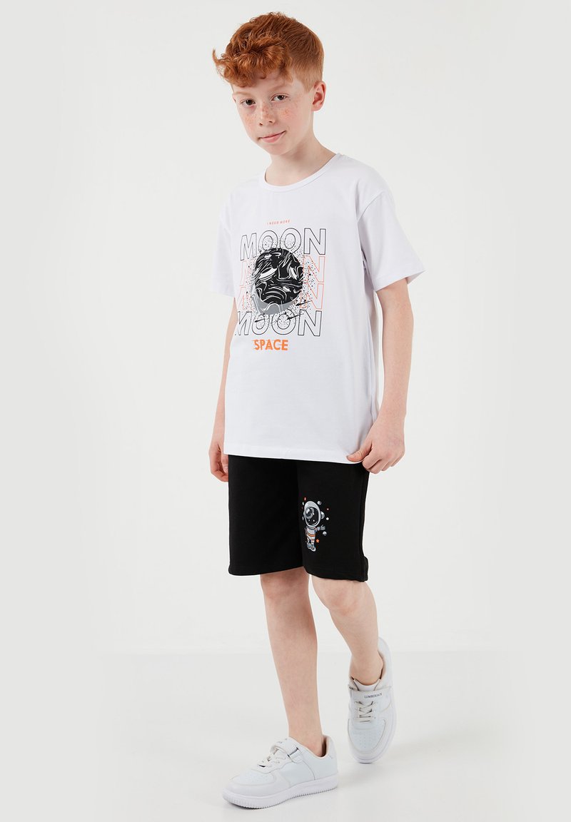 Garçon aux cheveux roux marchant, portant un t-shirt blanc avec l'inscription "Moon Space" et un design abstrait de lune, un short noir avec un graphique d'astronaute, et des baskets blanches.