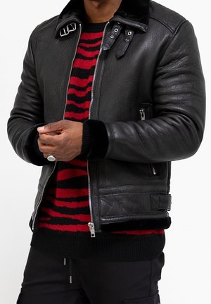 Mann trägt schwarze Lederjacke über rot-schwarz gestreiftem Pullover, hält die Hände zusammen und trägt einen silbernen Ring am linken kleinen Finger.