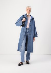 Cappotto lungo in denim con bottoni, colore azzurro chiaro, maniche ampie e spacchi laterali, abbinato a una maglietta rosa e jeans blu. Ballerine nere.