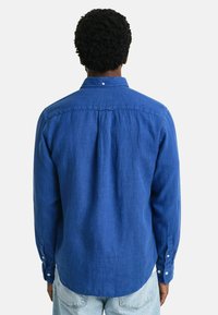 Camisa de lino azul con cuello abotonado, mangas largas y un pliegue en la parte trasera. Presenta botones blancos en los puños.