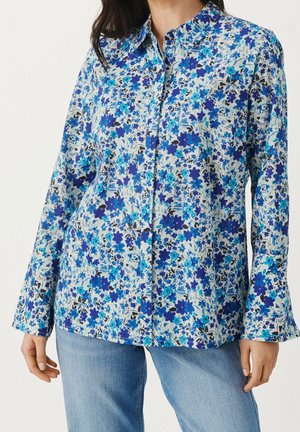 Mujer vistiendo una camisa de botón de manga larga con estampado floral azul, combinada con jeans de un azul claro.