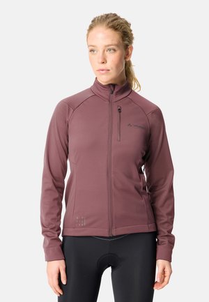 Vrouw met blond haar in een staart, gekleed in een mauve ritsvest en zwarte fietsbroek, staand tegen een effen achtergrond.