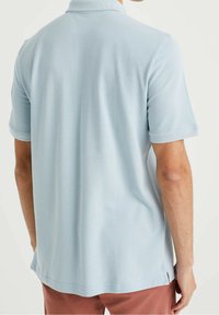 Homme portant un polo à manches courtes bleu clair et un pantalon couleur rouille, vu de dos contre un fond uni.