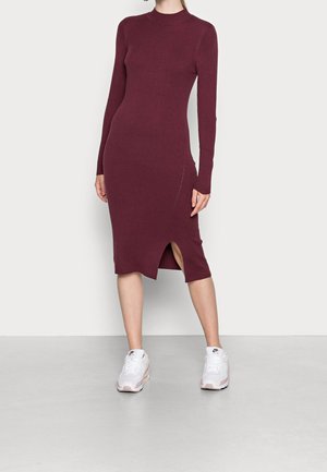 Kobieta ubrana w dopasowaną, bordową, dzianinową sukienkę midi z długim rękawem i rozcięciem z boku, w połączeniu z białymi sneakersami Nike, stojąca na szarej podłodze.