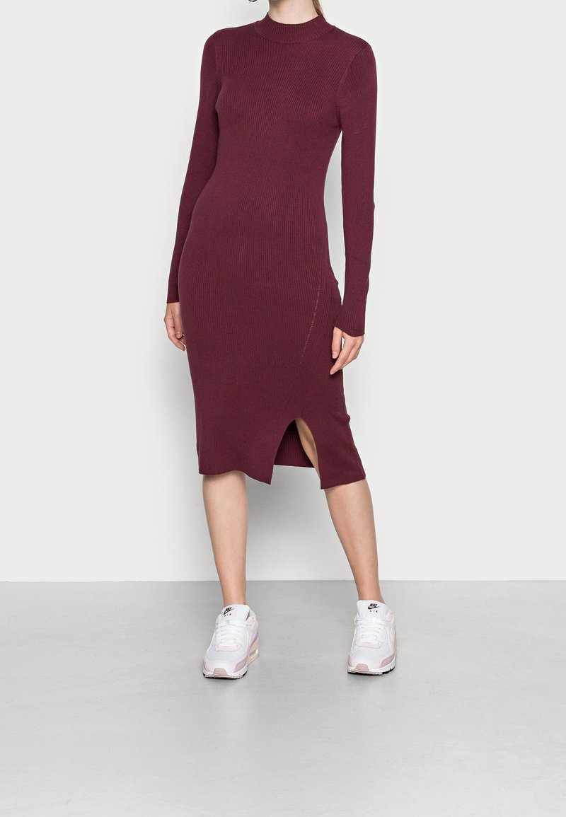 Femme portant une robe midi en maille bordeaux ajustée à manches longues avec une fente latérale, associée à des baskets blanches Nike, debout sur un sol gris.