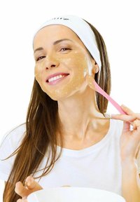 Kvinde med lyst brunt hår iført et hvidt pandebånd og skjorte, der påfører en gylden skrub på sit ansigt med en pink spatula. Hvid baggrund.