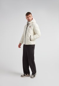 Vadderad vit jacka med huva, dragkedjestängning, logotypdetalj och svarta sidofickor. Stylerad med svarta byxor och sneakers.