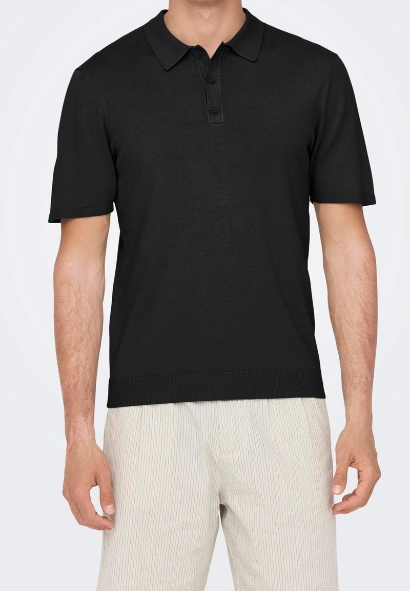 Homme portant un polo noir à manches courtes et un pantalon clair à rayures verticales, vu des épaules jusqu'à la mi-cuisse.