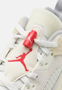 Baskets blanches avec un design perforé, dotées d'une étiquette de logo Jordan rouge et d'une silhouette brodée rouge sur la languette, maintenues par des lacets blancs.