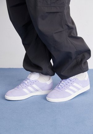 Person iført lyse lilla Adidas Gazelle sneakers med hvide striber, hvide sokker og løse sorte bukser, stående på blåt tæppe.
