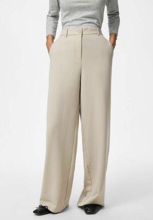 Pantalon beige à jambes larges avec fermeture bouton et zip porté avec un haut gris à manches longues et des chaussures noires pointues avec des nœuds.