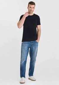 T-shirt nera in cotone con taschino sul petto, abbinata a jeans di denim blu. Il modello indossa scarpe da ginnastica bianche con suola marrone chiaro. Vestibilità e stile casual.