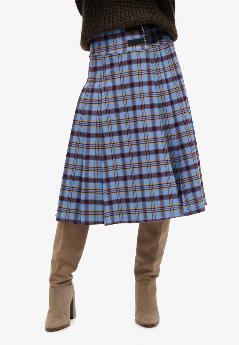 Jupe à carreaux gris-bleu au design plissé, avec des accents marron foncé et jaune, associée à des bottes en suède beige montantes.