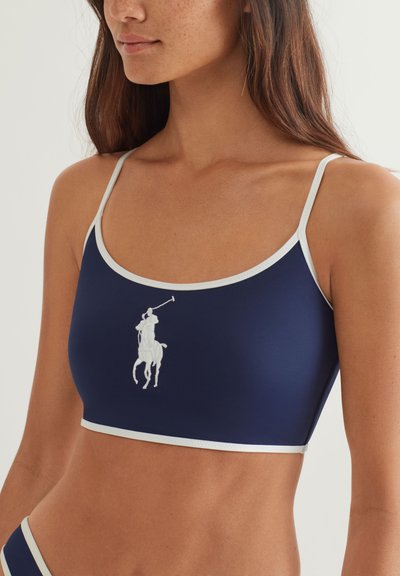 Γυναίκα που φοράει μπλε σκούρο κοντό τοπ με λεπτούς λευκούς ράντες και λευκό λογότυπο παίκτη πόλο στο κέντρο του στήθους.