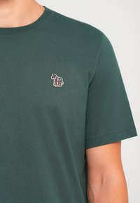 T-shirt verde scuro a maniche corte con un piccolo emblema di zebre ricamato e colorato sul lato sinistro del petto.