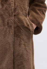Bruine faux bontjas met een zachte, gestructureerde afwerking, voorzien van zijzakken en een ontspannen pasvorm. De stof lijkt pluizig en warm.