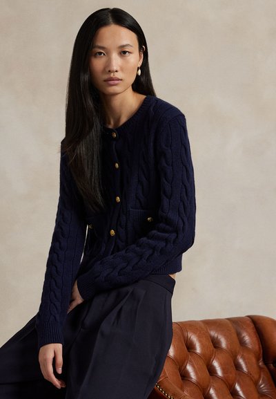 Kvinde med langt sort hår iført en kabelstrikket cardigan i marineblå med guldknapper og mørke bukser, sidder på en brun læderstol.
