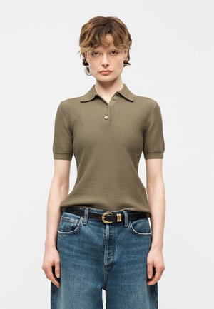 Fiatal személy olíva zöld rövid ujjú galléros pólót visel, amit kék magasderekú farmerral és fekete, arany csattal ellátott övvel visel.