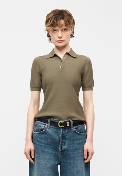 Jeune personne portant un polo vert olive à manches courtes, rentré dans un jean bleu taille haute avec une ceinture noire à boucle dorée.