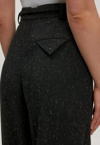 Close-up van zwart gespikkelde broek met een driehoekige knoopzak aan de achterkant, gedragen door een persoon met een wit T-shirt en zichtbare arm.
