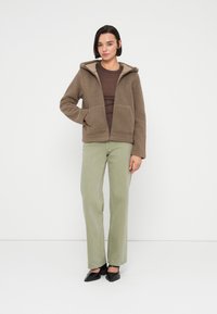 Καφέ φλις μπουφάν με κουκούλα και μπροστινές τσέπες, συνδυασμένο με ανοιχτό πράσινο παντελόνι ίσιας γραμμής και μαύρα παπούτσια με μυτερή μύτη.