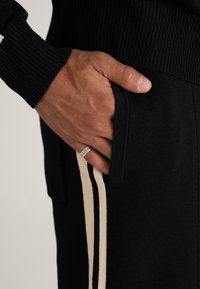 Man's hand with silver ring resting in black pants pocket with cream stripes, wearing a black ribbed sleeve sweater.