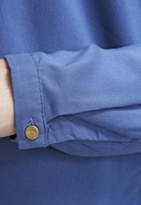 Blue, lightweight shirt fabric with a smooth texture. Features a buttoned cuff with a gold accent and stitching detail.
