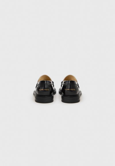 Svarta läderskor med rund tå och spännrem, med en tan-accent baktill och "FILLING PIECES"-logotyp i guld.