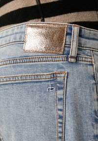 Vaqueros de denim azul claro con una etiqueta metálica dorada en la cintura y costuras naranjas. Incluye un logo bordado en blanco en el bolsillo trasero.