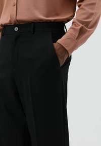 Zwarte, op maat gemaakte broek met een strak model, een gladde textuur en zijzakken. Gecombineerd met een lichtroze shirt met lange mouwen.