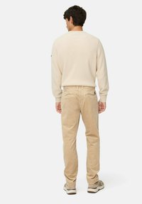 Man standing facing away wearing a cream ribbed sweater, beige corduroy pants, and beige sneakers against a plain white background.