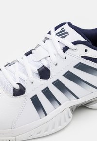 White athletic shoe featuring a textured leather upper, mesh tongue, navy accents, and gray branding stripes on the side.