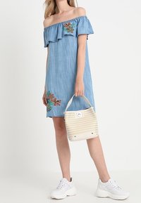 Denim mekko ilman olkaimia, jossa on röyhelöitä ja kukkakirjontaa punaisilla ja vihreillä sävyillä, jota täydentää valkoinen kudottu käsilaukku ja valkoiset lenkkarit.