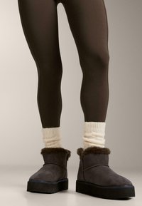 Noge, oblečene v temno rjave pajkice, kremno pletene nogavice in temno rjave gležnjarje z notranjo krzno podlogo ter debelim črnim podplatom na enostavni tleh.