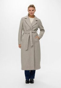 Cappotto di lana grigio chiaro con design avvolgente, ampie rever, cintura in vita e tasche laterali, abbinato a jeans scuri e stivali neri.