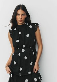 Schwarz gepunktetes Kleid mit Rüschenschultern und volantartigen Akzenten. Hergestellt aus leichtem Stoff, mit großen weißen Punkten und einer taillierten Taille.