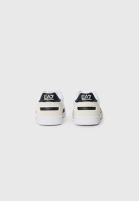 Sneakers brancos e bege com detalhes pretos. Apresentam uma textura suave, um colarinho acolchoado e um logótipo distinto na parte de trás. Sola de borracha.