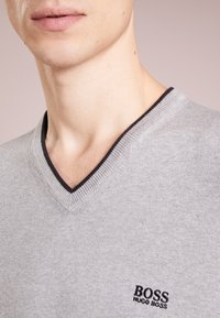 Grauer V-Ausschnitt-Pullover aus weichem Stoff, mit schwarzem Besatz am Ausschnitt und einem kleinen gestickten Logo auf der Brust.