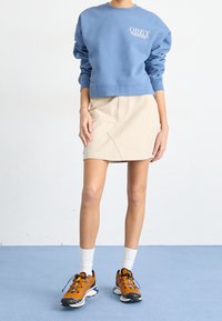 Blå sweatshirt med logotyp, beige kjol med fickor och orangea träningsskor med svarta detaljer och vita strumpor. Enkel, modern design.