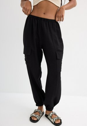 Pantalones cargo negros con cinturilla elástica, que cuentan con bolsillos laterales. Combinados con sandalias deslizantes de rayas en blanco y negro. Textura suave.
