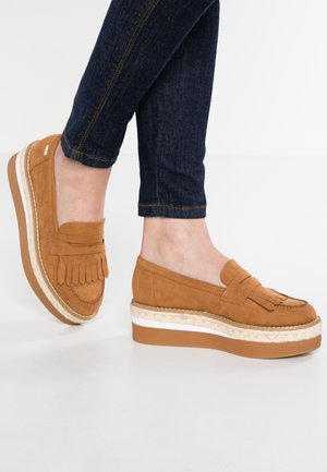 Fødder iført lysebrune ruskind platform loafers med kvaster, kombineret med mørkeblå opkrogede skinny jeans på hvid baggrund.