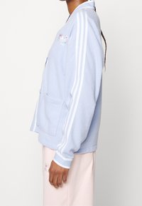 Light blue fleece cardigan with white three-stripe accents, buttoned front, and patch pockets, featuring floral embroidery on the chest.