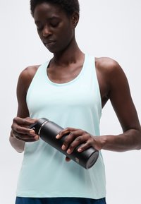 Femme en débardeur bleu clair tenant et ouvrant une bouteille d'eau isolante noire sur un fond blanc uni.