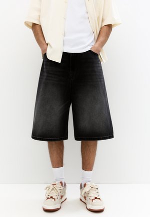 Personne portant un short en jean noir longueur genou, des chaussettes blanches, des baskets beige et marron, un t-shirt blanc et une chemise crème à manches courtes boutonnée.