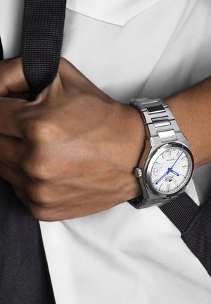 Montre en acier inoxydable argenté avec un cadran blanc texturé, aiguilles bleues et fermoir déployant. Portée sur une main tenant une sangle noire.